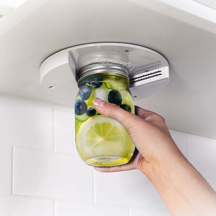Hand using a smart kitchen tool to open a jar with lemon and blueberry infused water under a cabinet.