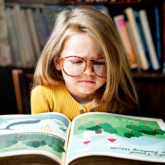 Young girl with large glasses reading a colorful book, reflecting concerns about aspects of modern life breaking down.