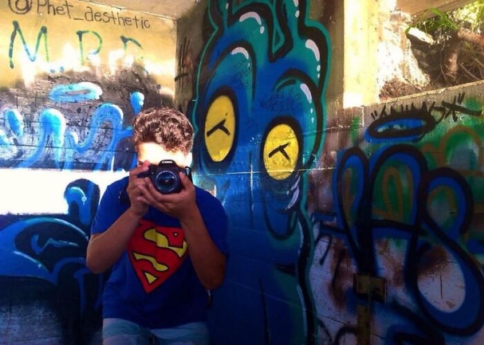 Person wearing a Superman shirt taking a photo in front of colorful graffiti in an urban setting, unsettling morbid knowledge theme.