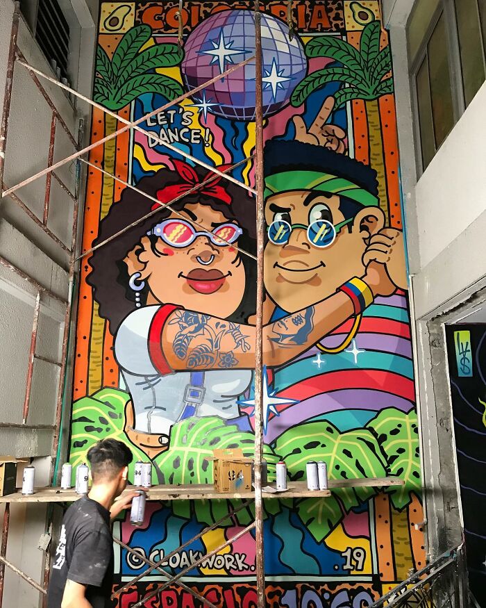 Vibrant street art mural of two dancing characters with colorful glasses and tropical background being painted on a large wall.