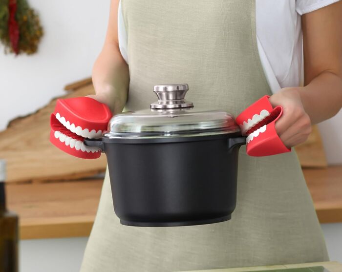 Person wearing clever kitchen problem-solver mitts shaped like teeth, holding a pot with a glass lid in a kitchen.