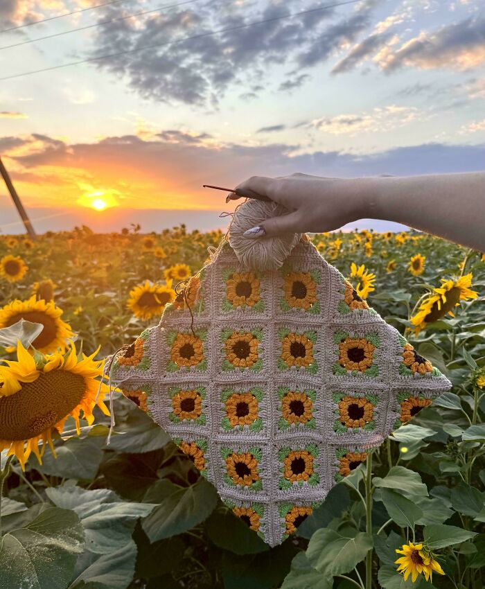 Hand holding a crochet piece with sunflower patterns in a sunflower field during a vibrant sunset, showcasing crochet enthusiasts' art.