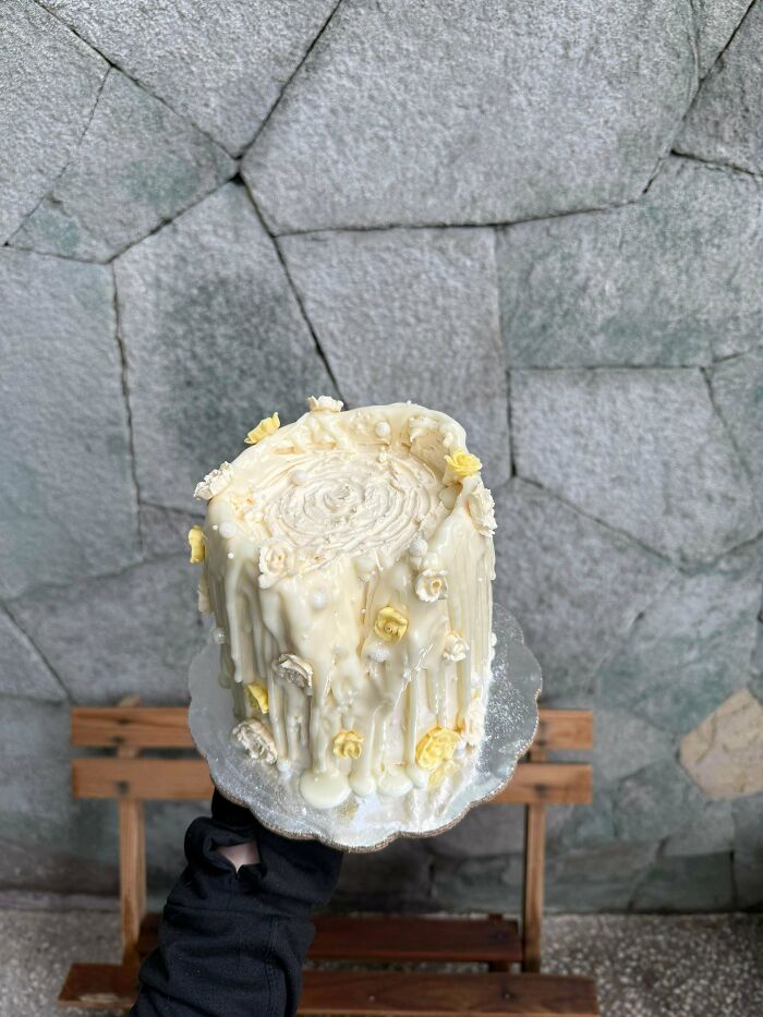 Hand holding a gorgeous baked goods cake with white frosting and yellow flower decorations against a stone wall background.