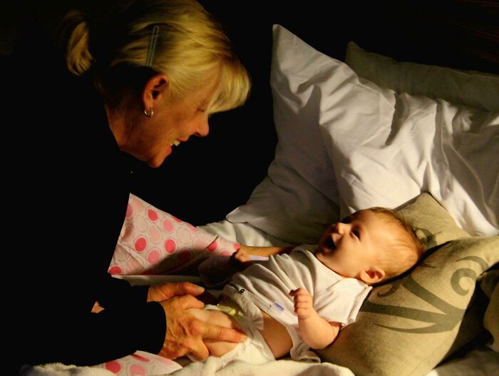 Woman smiling at a laughing baby on a bed, capturing a tender accidental renaissance moment with warm lighting.