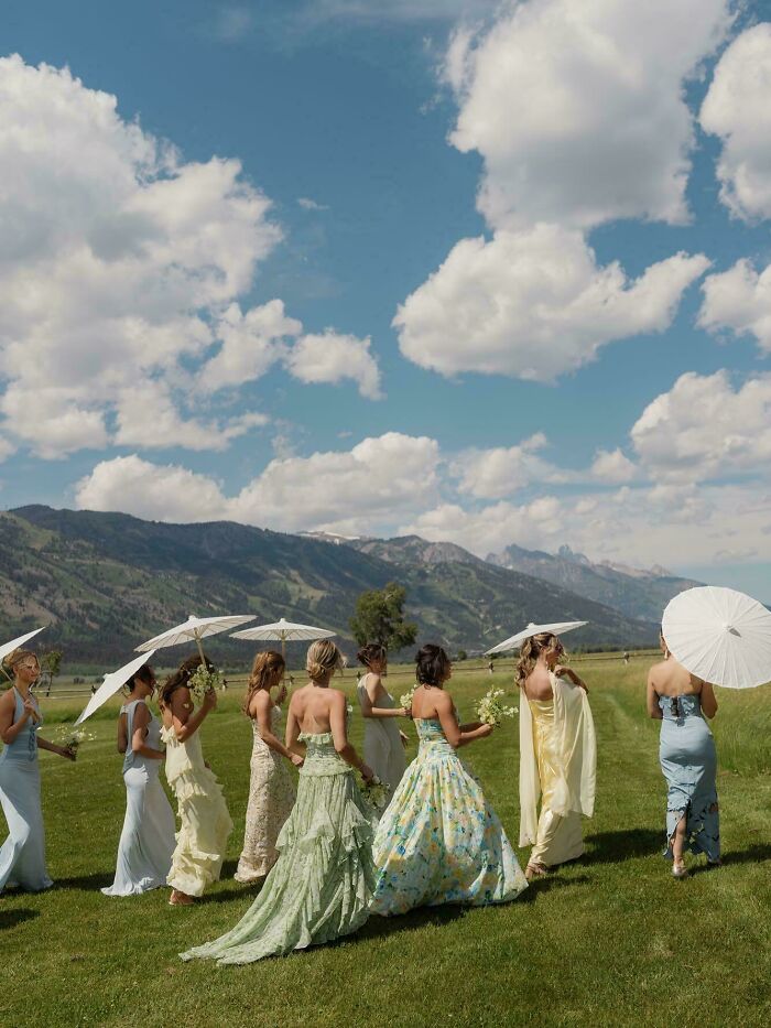 Bridesmaids in elegant dresses holding parasols, walking in a scenic landscape, capturing accidental renaissance photos.