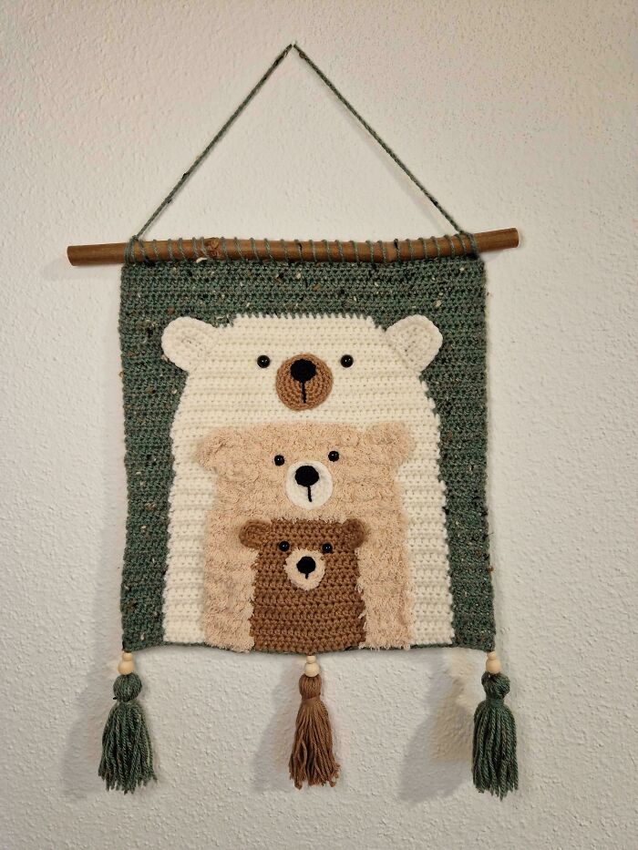 Crochet wall hanging featuring three nested bears in green, beige, and brown, with tassels on a wooden dowel.