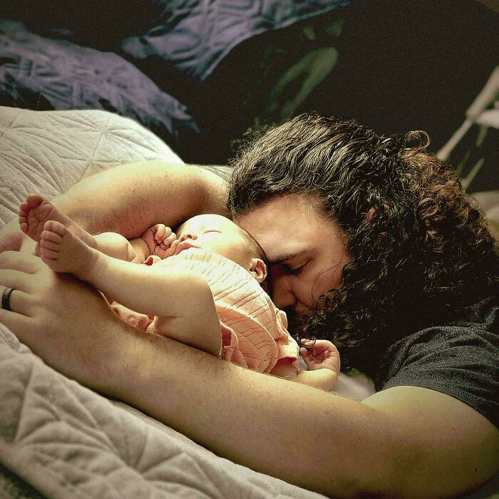 Father with curly hair tenderly cuddling a sleeping baby, creating a beautiful accidental Renaissance photo moment.