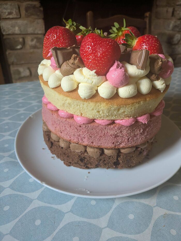 Three-layer baked goods cake with chocolate, pink and vanilla layers, topped with strawberries and cream dollops.