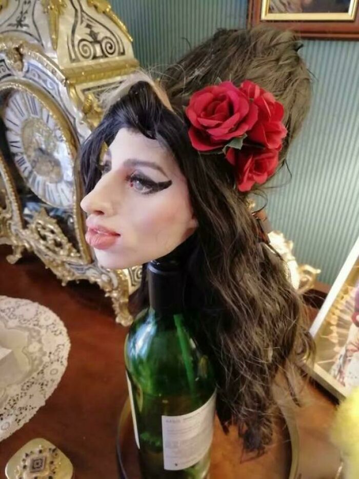 A wine bottle topped with a detailed mannequin head featuring dramatic makeup and red flowers, showing awful taste but great e*******n.