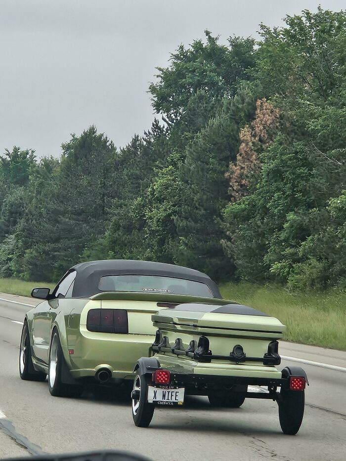 Green convertible Mustang towing matching coffin trailer with custom license plate on highway, example of awful taste but great e*******n