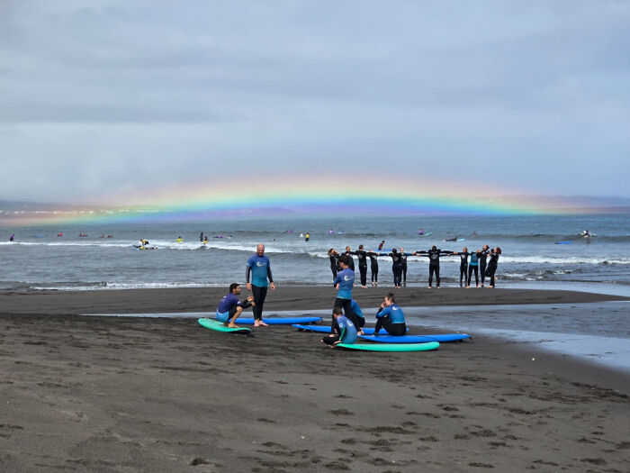 Group of people in wetsuits on a beach with surfboards and a vibrant rainbow over the ocean, moderately interesting scene.