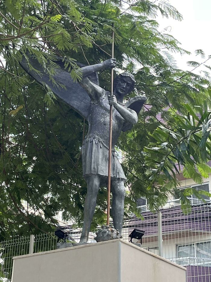 Statue of a winged figure holding a long spear in a park setting, an example of moderately interesting art shared by people.