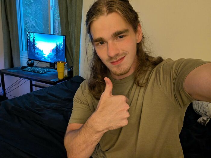 Young man with fierceflow beautiful hair giving a thumbs up, sitting on a bed in a casual room with a computer.