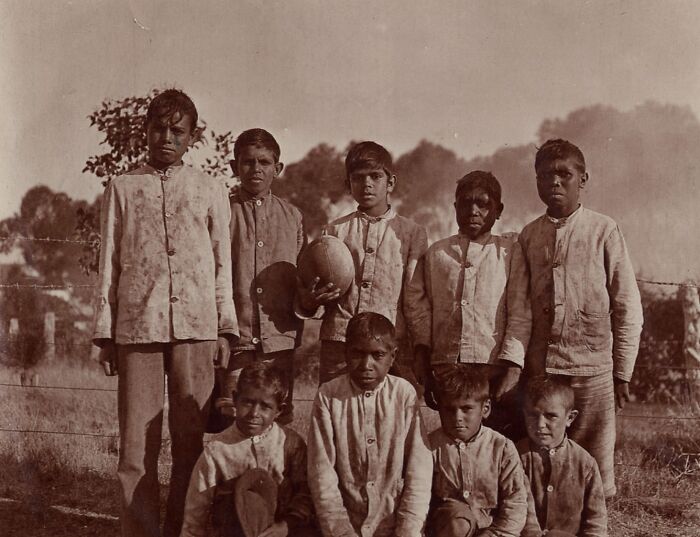 Group of Indigenous Australian children from the 1900s standing outdoors wearing similar clothes in a rare glimpse photo.
