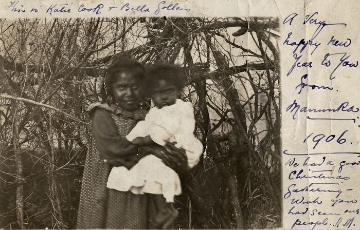 Two Indigenous Australian children in 1906, one holding the other, surrounded by dense branches in a rare historical photo.