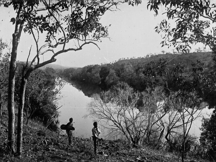 Two Indigenous Australians standing by a river in the 1900s surrounded by trees and natural landscape.