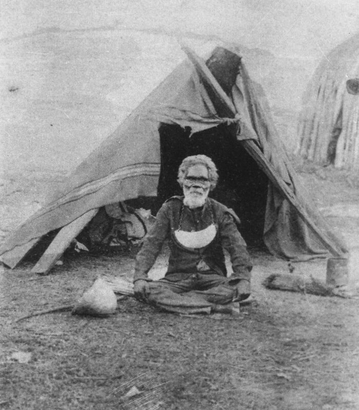 Elder Indigenous Australian man sitting cross-legged outside a traditional tent in early 1900s black and white photo.