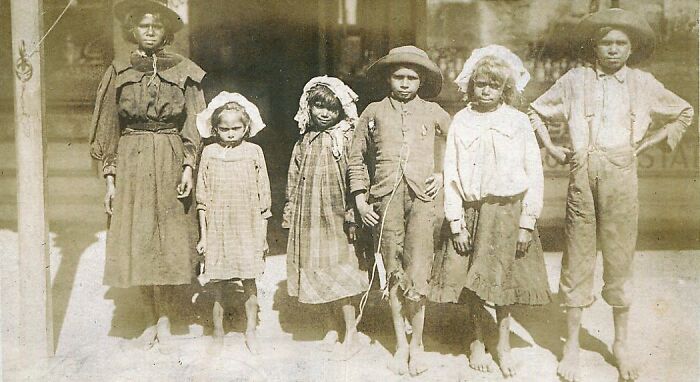 Group of Indigenous Australian children from the 1900s standing barefoot in worn clothing, offering rare glimpses into history.