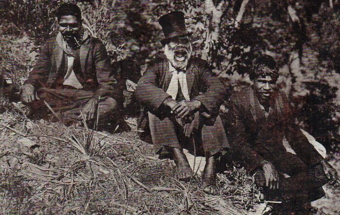 Three Indigenous Australian men from the 1900s sitting outdoors, capturing rare glimpses through striking historic photos.