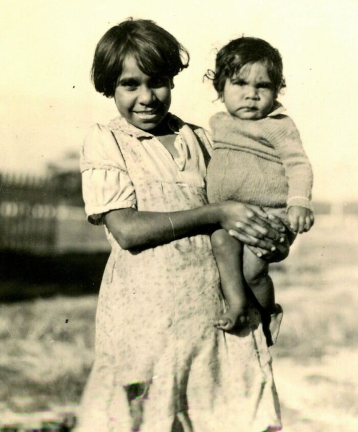 Young Indigenous Australian girl holding a toddler, capturing rare glimpses into 1900s Indigenous life in a black and white photo.