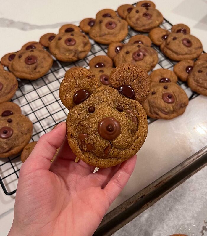 Close-up of a hand holding a bear-shaped chocolate chip cookie among other incredible bakes cooling on a rack.