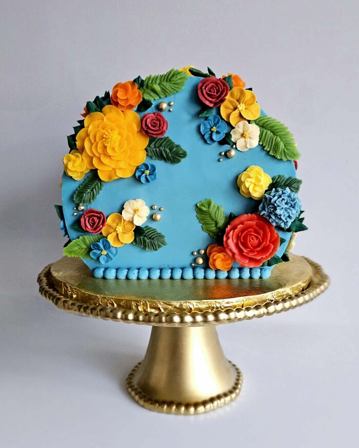 Blue frosted cake decorated with colorful buttercream flowers on a gold stand, an incredible bake worthy of the spotlight.