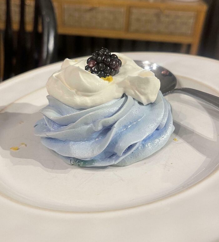 Light blue meringue bake topped with whipped cream and a blackberry on a white plate with a spoon.