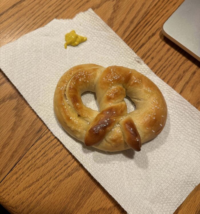 Soft golden baked pretzel with coarse salt on white napkin next to a small dollop of mustard on a wooden table.