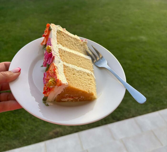 Slice of an incredible bake with floral icing decorations on a white plate held outdoors with a fork nearby