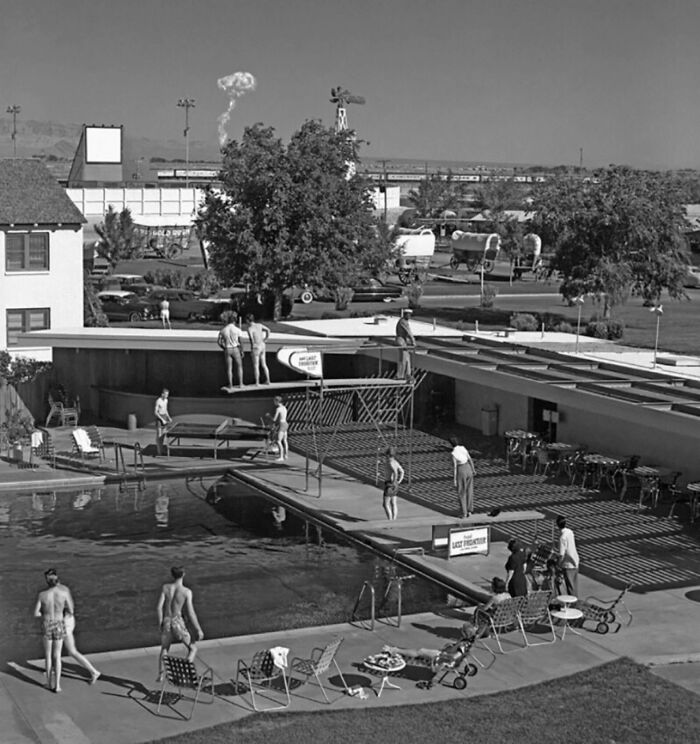 Black and white historical photo of people by a swimming pool with a train and wagons in the background.