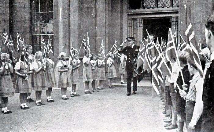 Schoolchildren holding flags greet a uniformed man outside a building, a historic scene of schools worldwide during World War II.