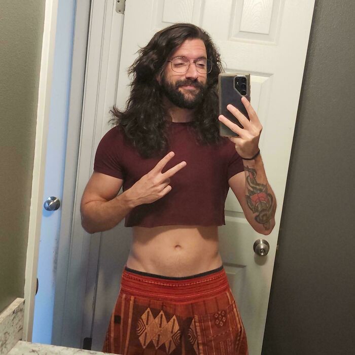 Man with fierceflow beautiful hair taking a mirror selfie wearing a cropped shirt and patterned pants, showing a peace sign.