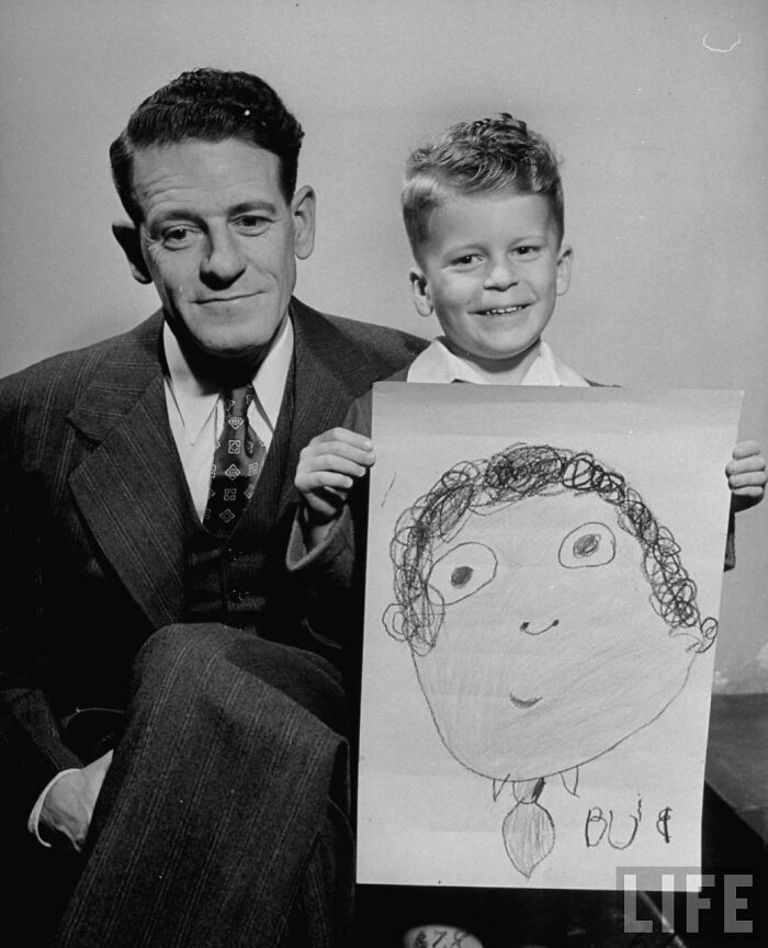 Young boy in 1949 holding a humorous kids drawing of his dad from memory, sitting next to the father in a suit.