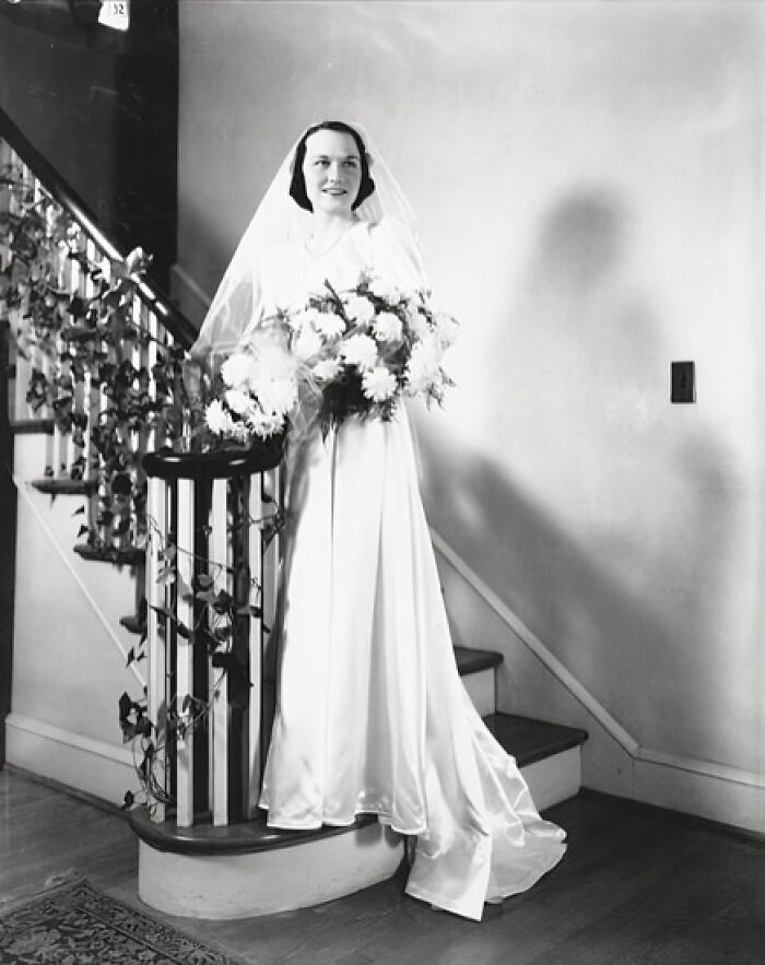 Bride in a 1940s wedding dress holding a bouquet, standing by a staircase decorated with vines during wartime wedding.