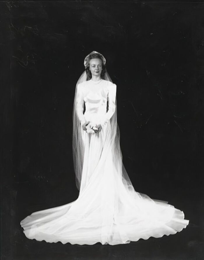 1940s wedding bride in long white gown and veil standing against a dark background, vintage wedding photo.