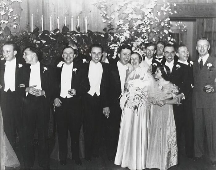 Black and white 1940s wedding photo showing a joyful bride and groom surrounded by smiling wedding party in formal attire.