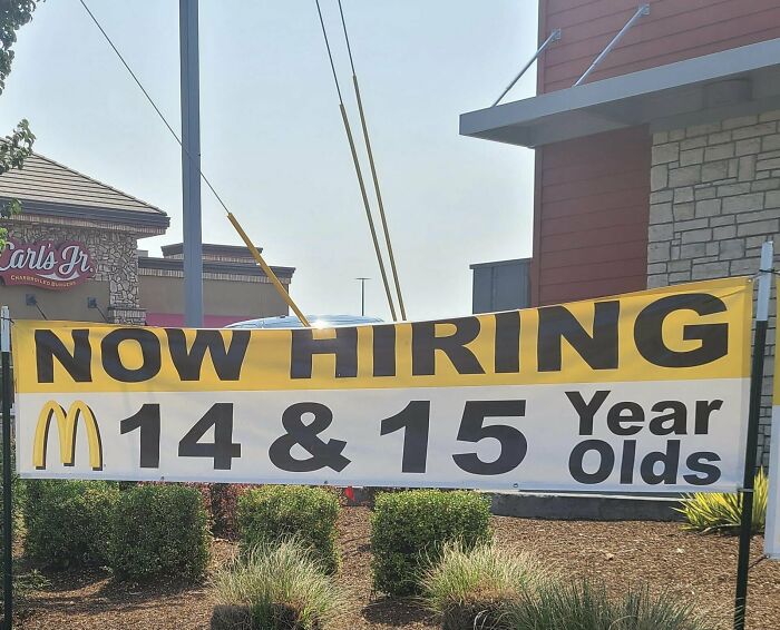 Hiring sign for 14 and 15 year olds outside a fast food restaurant, raising questions about labor and society changes.