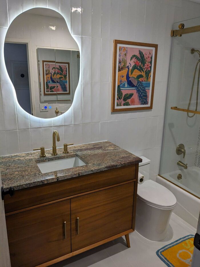 Modern bathroom corner with proud home decorating featuring wood vanity, granite countertop, illuminated mirror, and colorful peacock artwork.