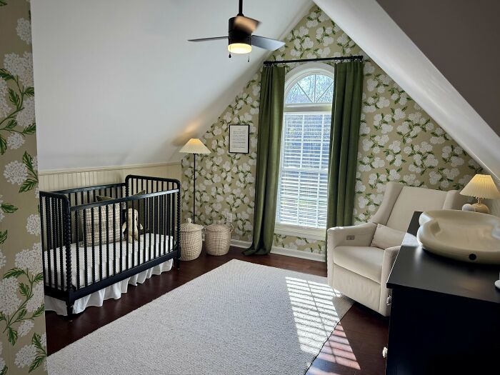 Cozy decorated nursery corner with floral wallpaper, black crib, white armchair, and natural light under sloped ceiling.
