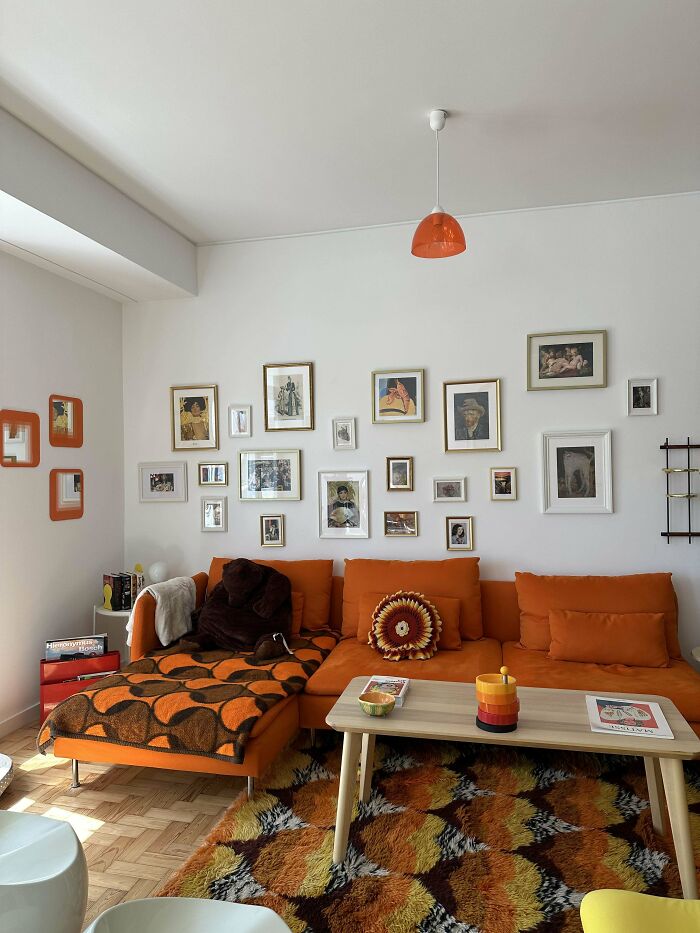 Cozy living room corner with orange couch, patterned throw, vintage art on walls, and colorful retro rug showcasing home decorating pride.