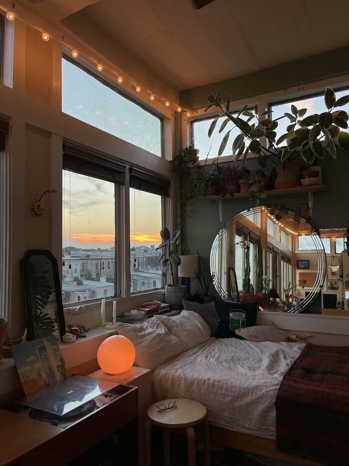 Cozy corner of a home decorated with hanging plants, a round mirror, warm lighting, and a city sunset view through large windows.