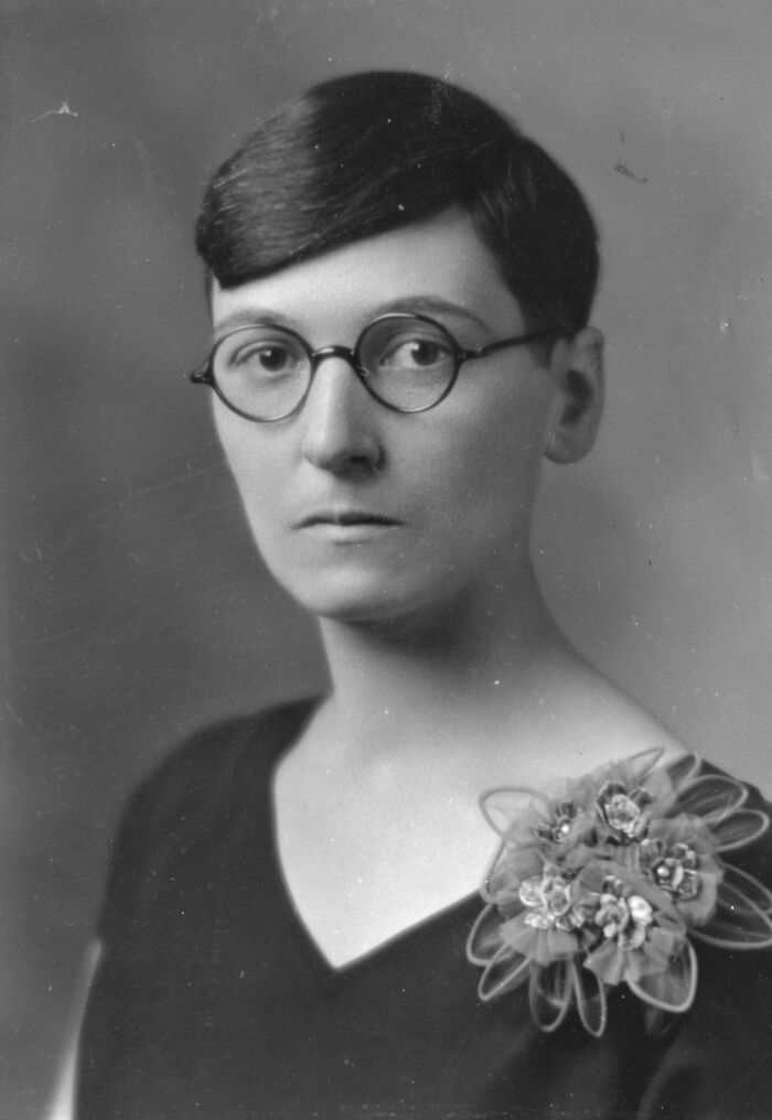 Black and white portrait of a woman scientist wearing round glasses and a dark top with floral decoration, vintage style.