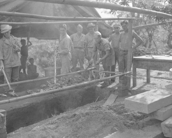WWII soldiers cooking food over an open fire trench while others watch in a jungle encampment during rare WWII moments.