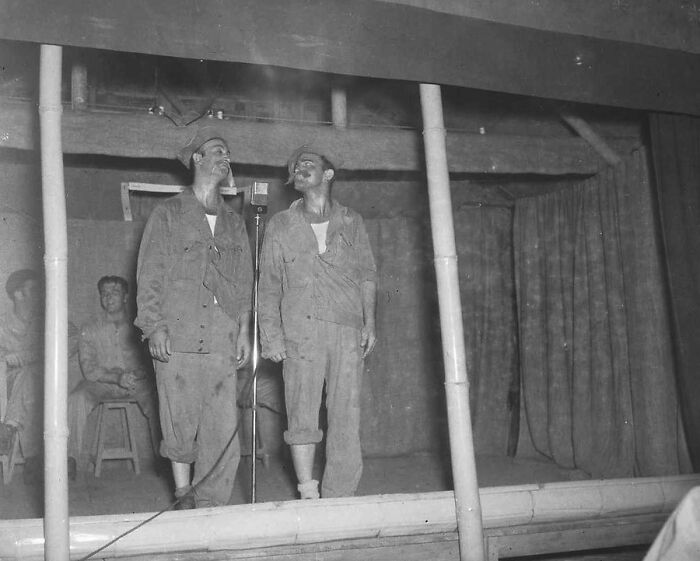 Two WWII soldiers smoking and standing near a microphone on a simple stage in a rare historic black and white photo.