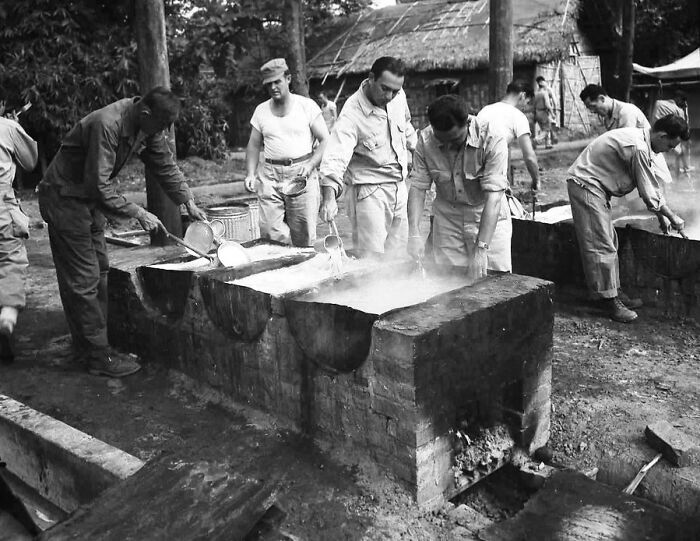 Black and white WWII photo showing soldiers cooking and washing at outdoor brick stoves in a camp setting.
