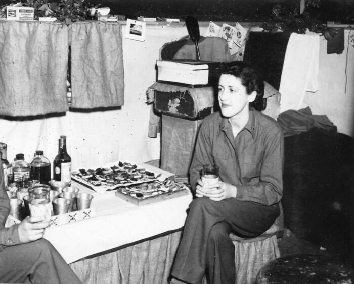 Rare WWII photo showing a woman soldier sitting beside a table with food and drinks in a wartime setting.