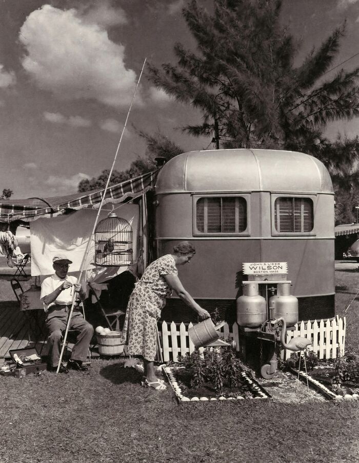 1950s Florida everyday life scene with a woman watering plants and a man fishing near a vintage trailer home.