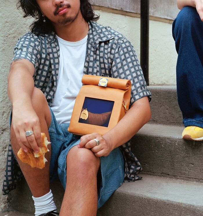Young man sitting on steps holding a croissant, wearing an unusual Nik Bentel accessory blending everyday objects with fashion.