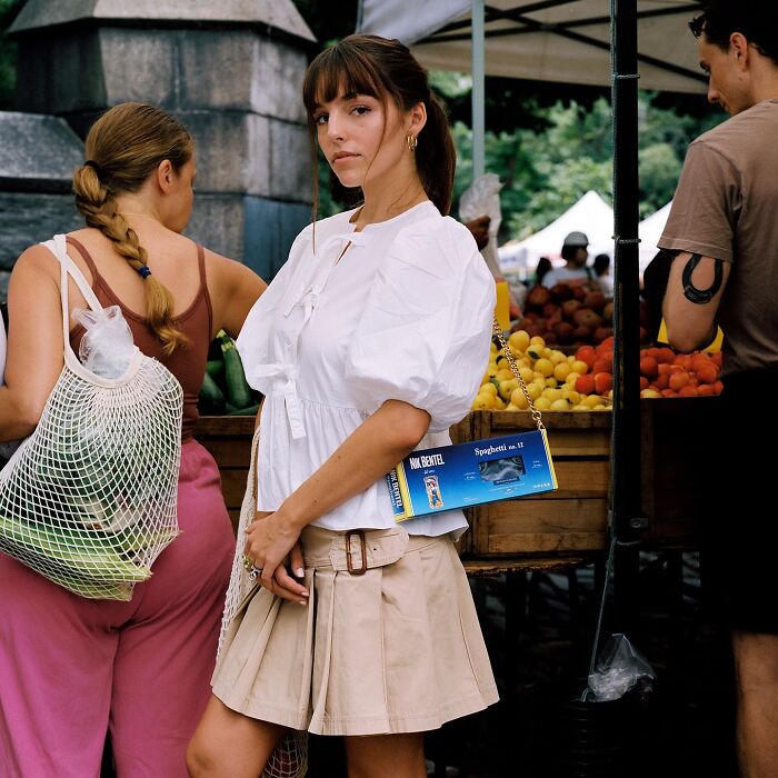 Woman at outdoor market wearing unusual accessories by Nik Bentel, including a handbag made from a spaghetti box.