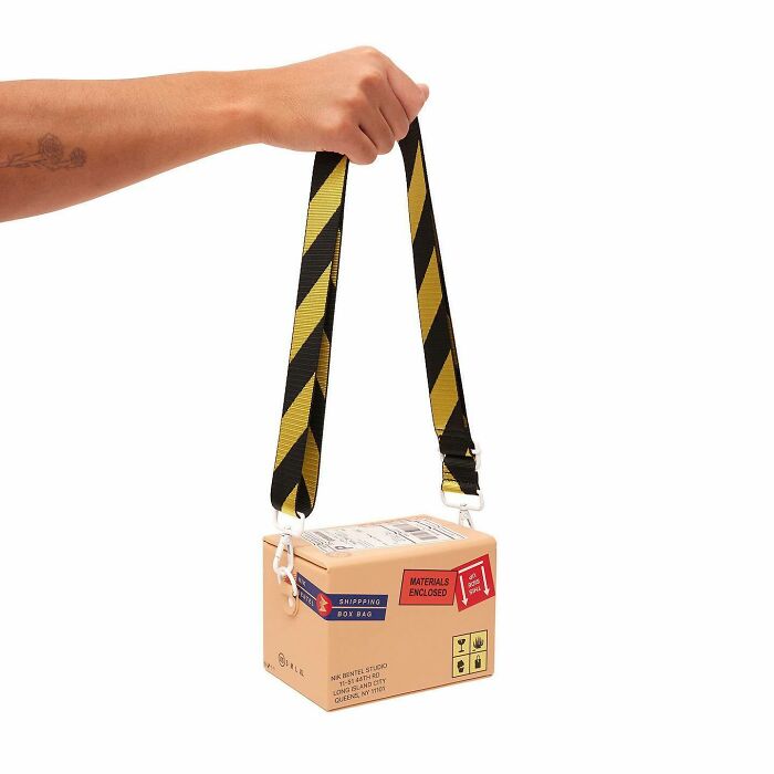 Hand holding a Nik Bentel unusual accessory shaped like a shipping box with a black and yellow striped strap.